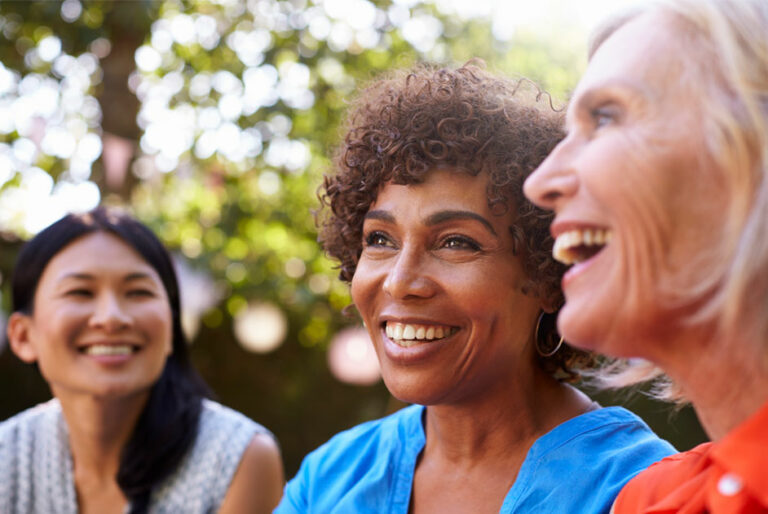 Três mulheres estão ao ar livre e sorrindo. Estão falando sobre saúde da mulher