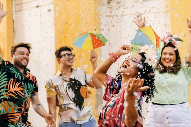 Grupo de amigos comemorando o Carnaval ao ar livre, jogando confetes no ar em clima de diversão.