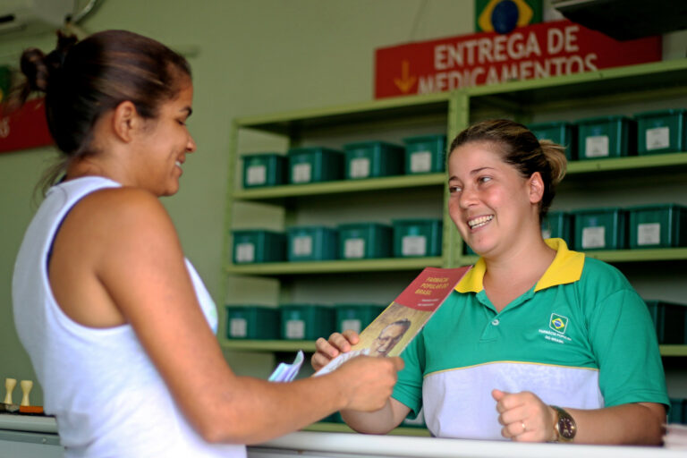 Uma mulher está no balcão de uma unidade da Farmácia Popular, onde é atendida por uma funcionária de uniforme verde e amarelo. Ambas sorriem. Ao fundo, há prateleiras com caixas e uma placa com os dizeres "ENTREGA DE MEDICAMENTOS".