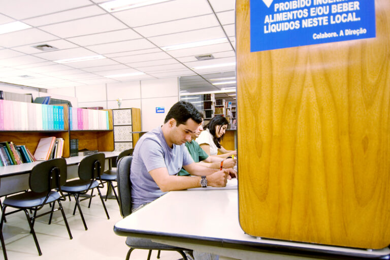 Dois homens e uma mulher trabalham atentamente, sentados lado a lado.. Atrás deles, uma bancada com livros e cadeiras vazias. Ao fundo, estantes com livros