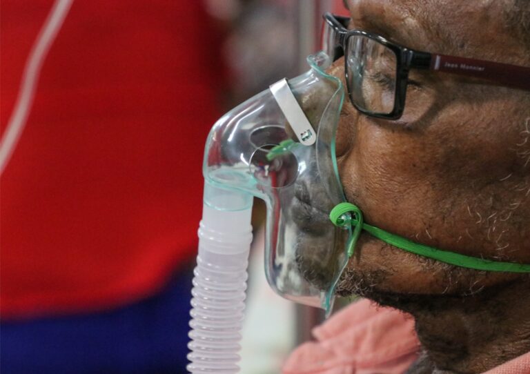 imagem em close de um homem de óculos, com uma máscara de oxigênio no rosto, como quem está em inalação, por doenças respiratórias, em um ambiente que parece ser uma unidade de saúde.